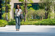 © Jirakul - young woman with curly hair walks outdoors, smiling while using her smartphone. She wears striped sweater and wide leg jeans, surrounded by greenery in modern urban setting