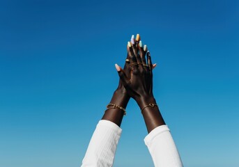 black woman's hands adorned with golden jewelry, pastel painted nails intertwining against bright bl