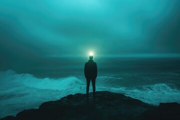 Wall Mural - Silhouette of a man standing on a cliff overlooking a teal ocean, under a bright light.