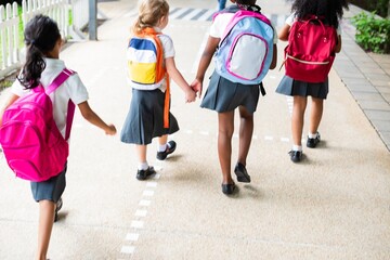 Poster - Four young girls with backpacks walk together on a path. School uniforms, backpacks, and friendship are highlighted in this cheerful scene. Cute little kids with backpacks in school.