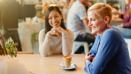 Poster - Two women, one Asian and one Caucasian, enjoy coffee together in a cozy cafe. The Asian woman smiles while the Caucasian woman sips her coffee, creating a warm, friendly and lively atmosphere.