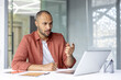 © Liubomir - Serious and focused man talking to colleagues and partners sitting at desk, using laptop for video call. Businessman inside office at workplace.