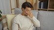 © Krakenimages.com - Young man sitting on a sofa in a living room covering his eyes with his hand, suggesting a moment of relaxation or contemplation in a cozy indoor setting.