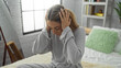 © Krakenimages.com - Woman sitting stressed on a bed in a cozy bedroom with hands on head, embodying stress and contemplation, surrounded by soft pillows and muted natural light from a large window.