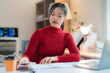 © Tj - Young Asian businesswoman wearing red turtleneck sweater working from her home office taking notes on a notebook and using her laptop, business and finance concept