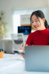 Fototapete - Young Asian businesswoman using smartphone and working with laptop while sitting at table with notebook and takeaway coffee cup at workplace in modern office