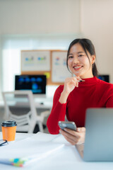 Fototapete - Young Asian businesswoman holding a smartphone and working with laptop while sitting at her desk in modern office, smiling and looking satisfied