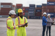 © ultramansk - Two logistics workers in high visibility safety gear and hard hats discuss operations at a busy shipping container yard. One is holding a tablet while pointing towards the cargo stacks.
