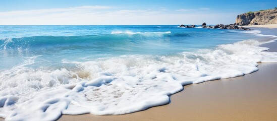 Aufkleber - Serene beach scene with soft waves rolling onto golden sand under a clear blue sky, cliff to the right, vibrant azure ocean in the background.