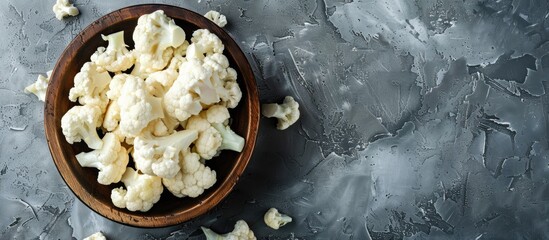 Poster - Freshly cut cauliflower florets in a round wooden bowl on a textured gray surface with scattered florets and ample blank space on the right