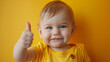 © Duka Mer - A cute happy smiling toddler baby in a yellow t-shirt shows a thumbs up gesture on a colored background.