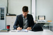 © Nuttapong punna - Confident businessman working on finance analysis with a tablet and laptop at office desk.  professional workplace concepts..