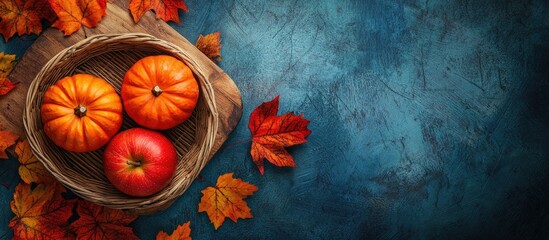 Poster - Vibrant autumn colors with orange pumpkins and a red apple in a woven basket surrounded by colorful leaves on a textured blue background