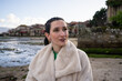 © asier - Young woman wearing braided hair and faux fur coat wandering through historic combarro, experiencing traditional stone architecture and peaceful waterfront of galician coastal village