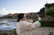 © asier - Young woman with braided hair photographing scenic combarro waterfront, wearing trendy coat while capturing traditional stone structures along coastal galician landscape