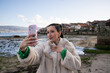 © asier - Young woman with braided hair wearing faux fur coat capturing selfie near traditional stone granaries of combarro, overlooking pontevedra estuary's scenic waterscape