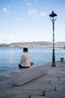 © asier - Tourist sitting on concrete bench, overlooking tranquil waters and distant hills of combarro, absorbing serene coastal landscape of galician seaside promenade