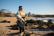 © asier - Female musician performing acoustic guitar, wearing fur coat and boots, standing on rocky beach during sunset with crashing ocean waves at punta faxilda