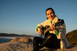 © asier - Female musician playing acoustic guitar, perched on rocky shoreline during golden sunset, capturing serene coastal moment