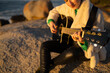 © asier - Female musician strumming acoustic guitar while perched on rocky shoreline, golden sunset rays illuminating peaceful coastal landscape
