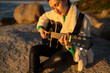 © asier - Female guitarist performing acoustic melody, sitting on seaside rock during golden sunset at punta faxilda
