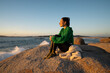 © asier - Young woman perched on seaside rock at punta faxilda, basking in golden sunset glow, gentle waves lapping rocky coastline, peaceful coastal scene unfolding
