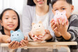 © Rakchanok - Asian family teaching children saving in piggybank, practicing finance planning for future, mother put coins in piggy bank and encourage children to be disciplined to save money left from school