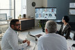 © pressmaster - High angle view at group of diverse medical experts with Middle Eastern male doctor participating in online medical conference at meeting table in clinic
