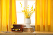 © Pixel-Shot - Retro radio receiver with headphones and flowers on chest of drawers near yellow curtains in room