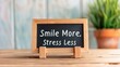 © Katawut - A wooden sign displaying the phrase 'Smile More, Stress Less' placed on a table near a plant, promoting positivity and mindfulness.