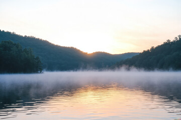 Wall Mural - Serene misty lake at dawn, surrounded by lush green hills and reflecting the early morning sky.
