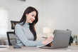 © PRIME STOCK LAB - Asian Businesswoman Working at Desk With Laptop, Writing Notes in Notebook, and Smiling in Bright Office.