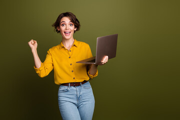 Poster - Young happy woman in trendy outfit celebrating success while holding a laptop, against a modern khaki background