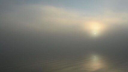 Wall Mural - Minimal landscape at calm foggy lake as sun rises behind a thick fog. Reflection in rippling water.
