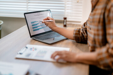 Poster - Businesswoman pointing at laptop screen showing trending product data while analyzing paperwork