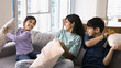 © fizkes - Young mother engaged in cushion fight with their two little children, having fun, holding cushions beating each other seated together on couch in living room. Active weekend games with kids at home