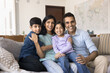 © fizkes - Indian couple and their two cute kids relaxing seated on couch looking at camera with joyful expression, feeling unconditional love, having harmonic relationships. Well-being loving family portrait
