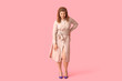 © Pixel-Shot - Young woman with curly hair posing in stylish beige shirt dress and purple heels on pink background, she poses with one hand on her hip, looking directly at the camera