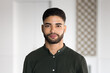 © fizkes - Young Arab man dressed in casual shirt standing indoors with serious expression, stare at camera, looks confident, head shot, office portrait. Human resources, employment, profile picture of applicant