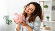 © Kunut - Young woman holding piggy bank, smiling joyfully in bright room