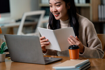 Aufkleber - Asian businesswoman reviewing notes on spiral notebook during video conference with laptop at workplace