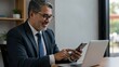 © Qbertstudio - Hispanic businessman using smartphone in office setting with laptop Represents modern work culture and technology integration