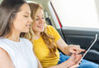 © Denis Rozhnovsky - Two smiling women sitting in a car are using a tablet, seemingly engaged in navigation or planning during a road trip.