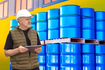 Poster - Pallets with barrels near man. Guy owner of oil refinery. Man with electronic tablet near barrels. Oil tanks behind worker. Specialist in helmet works at petroleum warehouse. Oil refinery contractor