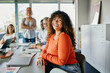 © Flamingo Images - Confident woman smiling in a modern office meeting setting. She looks directly into the camera, smiling, while her happy diverse colleagues sit behind her.