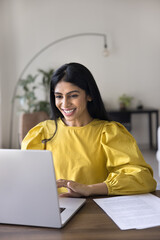 happy indian woman sit at table looks at laptop screen, read pleasant notice or news, get business o