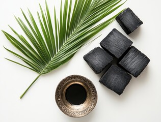 Poster - Elegant Arrangement of Charcoal Blocks with a Palm Leaf and a Silver Bowl of Ink on a Light Background for Artistic and Culinary Use