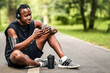 © Prostock-studio - Tired african sportsman sitting on park path, drinking protein and using smartphone, empty space