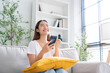 © Monster Ztudio - Happy young asian woman relax on comfortable couch at home texting messaging on smartphone, smiling girl use cellphone, chatting online message, shopping online from home