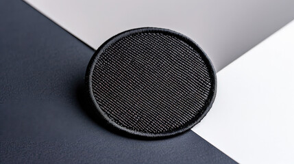 Poster - black circular fabric patch with finely woven texture is placed on contrasting black and white background, highlighting its detailed craftsmanship and sleek design